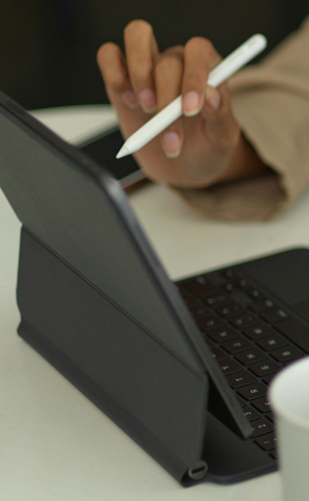 Tablet In Use A hand using a stylus on a tablet beside a cup and keyboard.